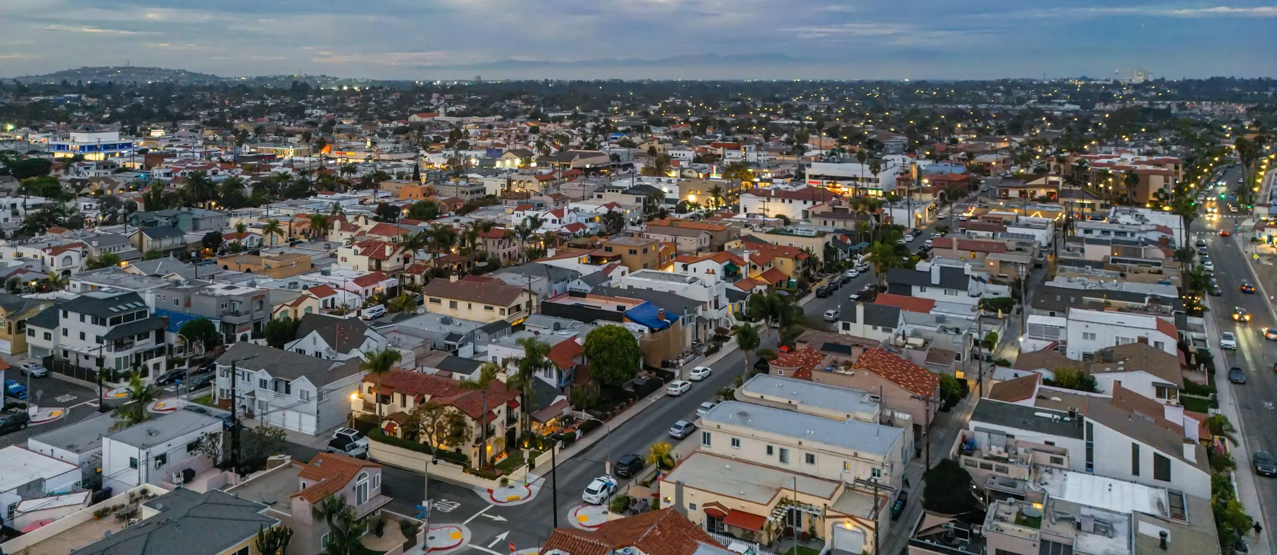 A Southern California neighborhood at dusk, seen from the air, porch lights and streetlights coming on across the rooftops